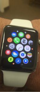 A white apple watch is pictured on a man's wrist. Many multi-colored apps are visible on the watch's home screen. 