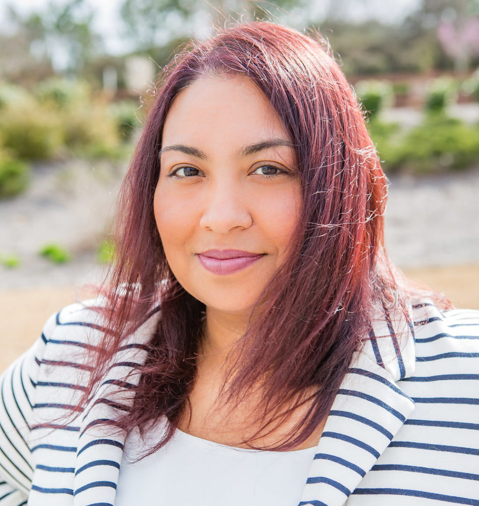 Elysia, a woman with auburn hair and brown eyes, wears a white shirt and blue and white jacket.