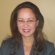 Tina, a woman with brown hair, wears wire-rimmed glasses, a grey blouse and jacket and is smiling. 