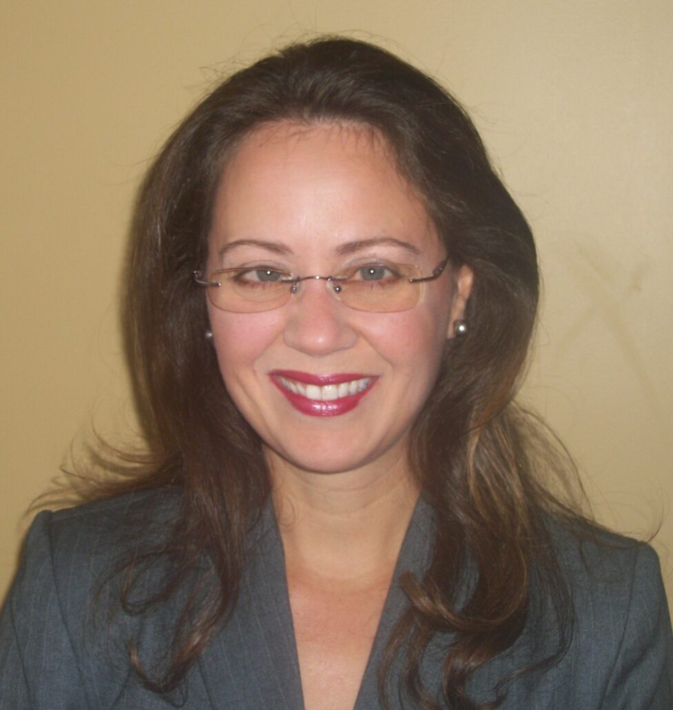 Tina, a woman with brown hair, wears wire-rimmed glasses, a grey blouse and jacket and is smiling.