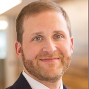 Nick, a white man with light brown hair and a beard, wears a white shirt, tie and black suit.