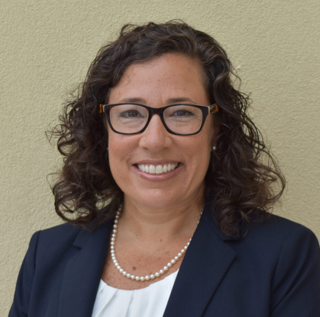 Sylvia, a woman with curly brown hair and glasses, wears a white blouse and dark blue jacket.