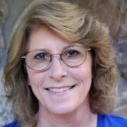 Peggy Pardue is a white woman with light brown, shoulder-length hair. she is wearing wire-rimmed glasses and a bright blue blouse. She is pictured smiling.