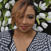 Elysia, a woman with auburn hair and brown eyes, wears a white shirt and blue and white jacket.