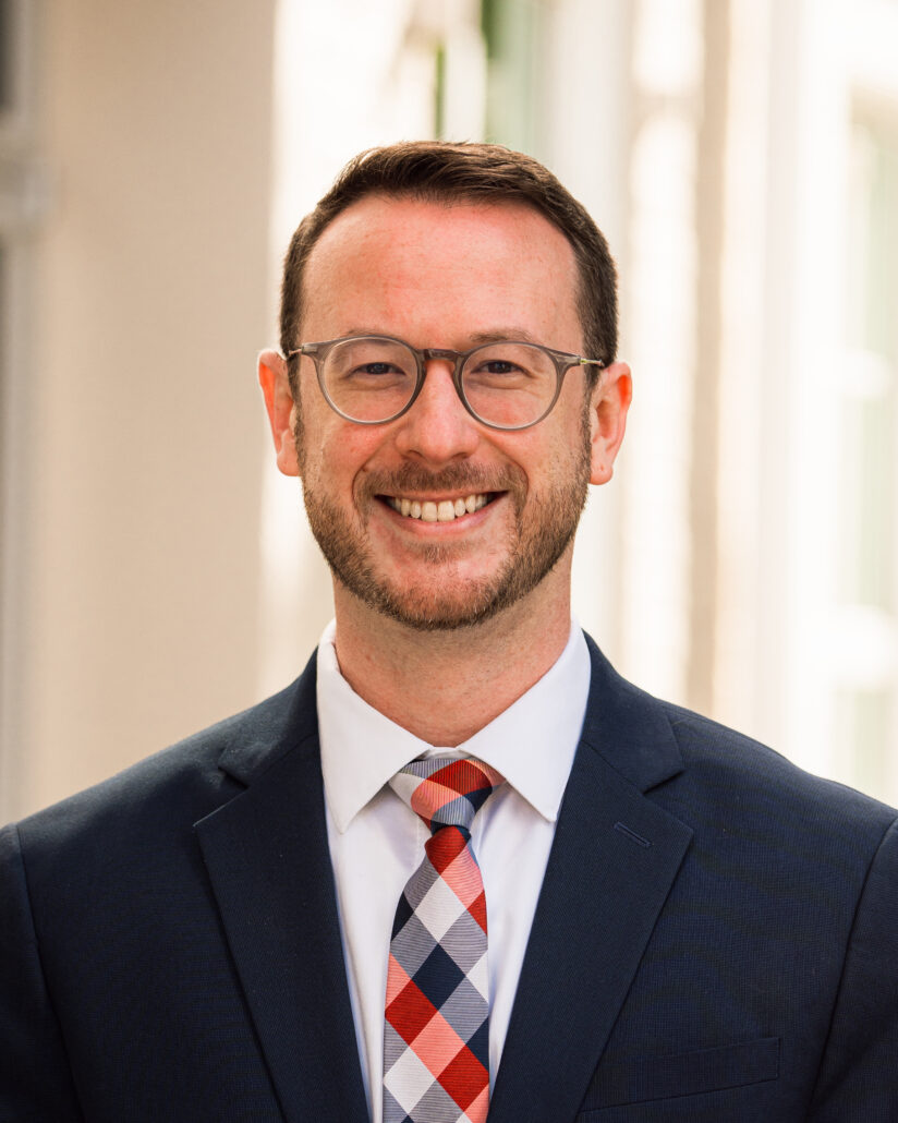 Drew, a white man with brown hair and a beard, wears a white shirt, plaid tie, and black suit.