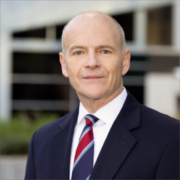 Chris, a white man with grey hair, wears a white shirt; red, navy, and white striped tie; and black suit. 