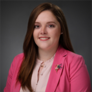 Kayla, a woman with dark brown hair, wears a pale pink blouse, bright pink jacket, and gold fairy pin on the lapel.