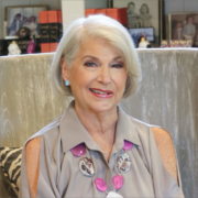 Nancy, a white woman with white hair, wears a pale grey blouse with cut out sleeves and pink and white embroidery.