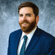 Kris, a white man with brown hair and a beard, wears a white shirt, teal tie, and navy blazer.