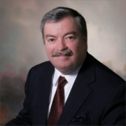 Mark, a white man with grey hair, wears a white shirt, burgundy tie, and black blazer. 