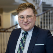 S. Collins Saint is a person with short, golden-brown hair. They wear a pale blue button down shirt, a navy and green striped tie, a navy suit, and brown circular glasses.