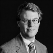 Dan, a white man with blond hair, wears a white shirt, plaid tie, and grey jacket, and round wire-framed glasses. The photo is in black and white.