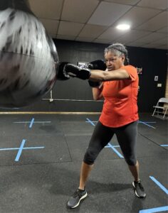 Tamesa, a Black woman with grey hair, is pictured wearing boxing gloves and punching a boxing bag. She wears an orange shirt and black pants.