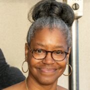 Tamesa, a Black woman with grey hair, wears brown glasses. Her hair is in a bun, and she is smiling.