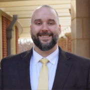 Benjamin, a white man with a brown beard, wears a white shirt, gold tie and black jacket. He stands with a brick building behind him.