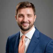 John, a white man with brown hair and a beard, wears a white shirt, peach tie, and navy suit.