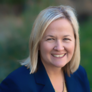 Patti, a white woman with shoulder-length blond hair, wears a blue suit. In the background is blurred foliage. 