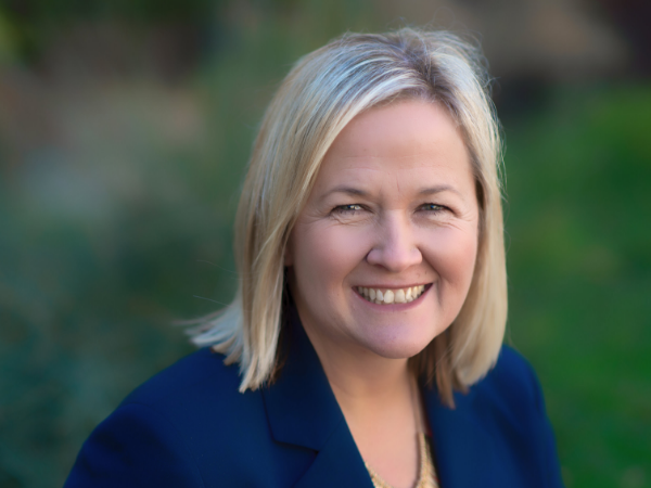 Patti, a white woman with shoulder-length blond hair, wears a blue suit. In the background is blurred foliage.