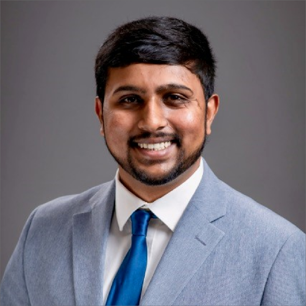 Surya, a man with black hair, wears a white shirt, pale grey suit, and teal tie.