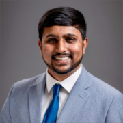 Surya, a man with black hair, wears a white shirt, pale grey suit, and teal tie. 