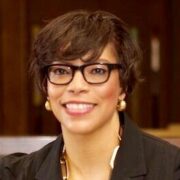 Stacey, an African American woman with short brown hair, wears brown glasses, gold earrings and a necklace, and a black suit.