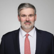 Evan, a white man with grey hair, wears a white shirt, red tie and black jacket. 