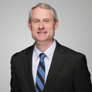 Paul, a white man with grey hair, wears a white shirt, black and blue tie, and black jacket.