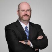 Thomas, a white man with a brown beard, wears a white shirt, blue striped tie, and black jacket.