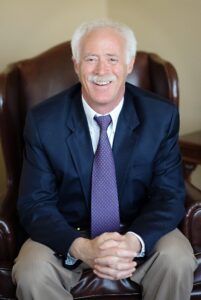 Kerry, a white man with white hair and a mustache, wears a white shirt, purple tie, navy jacket and khaki pants.