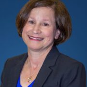 Rona, a woman with brown hair, wears a bright blue blouse and grey jacket. 