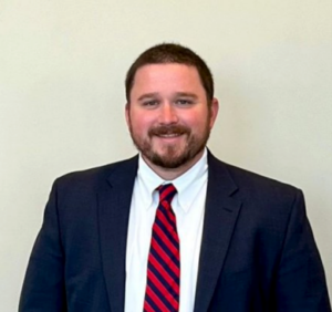 Tripp, a white man with brown hair, wears a white shirt, red and navy striped tie, and navy suit.