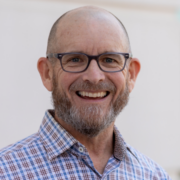 Will, a white man with a brown beard, and grey glasses, wears a blue and peach plaid shirt. 