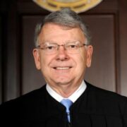 Bob Hunter, a white man with grey hair and wire-rimmed glasses, wears a white button-down shirt and pale blue tie and a black judge's robe.
