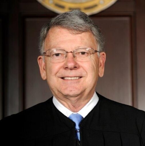 Bob Hunter, a white man with grey hair and wire-rimmed glasses, wears a white button-down shirt and pale blue tie and a black judge's robe.