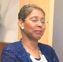 Kimberly, a Black woman with black hair and clear glasses, wears a blue blouse and jacket with sparkles and a white beaded necklace.