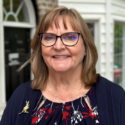 Leslee, a white woman with light brown hair and dark brown rimmed glasses, wears a navy dress with red and white flowers, a navy sweater, and a gold kangaroo pin. 