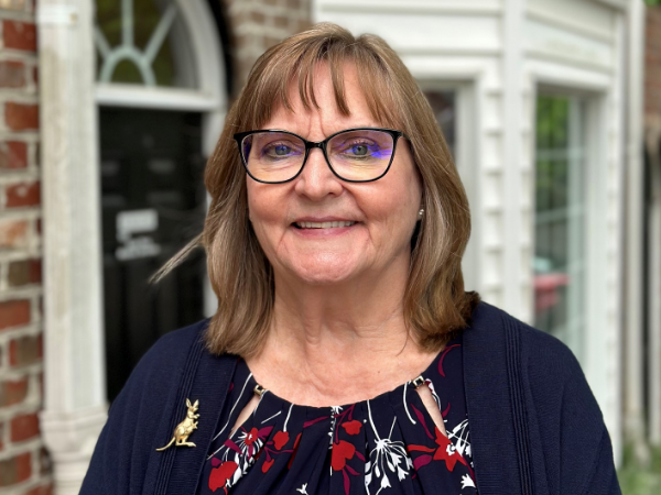 Leslee, a white woman with light brown hair and dark brown rimmed glasses, wears a navy dress with red and white flowers, a navy sweater, and a gold kangaroo pin.
