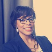 Stacey, a Black woman with black hair, wears black-rimmed glasses, a white blouse, black jacket and gold jewelry.