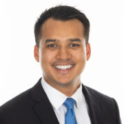 Ash, a man with black hair, wears a white shirt, bright blue tie, and black jacket, and he is smiling.
