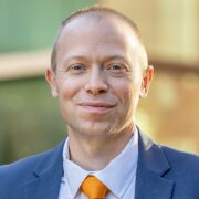 Matt, a man with light brown hair, wears a pale blue shirt, orange tie, and blue jacket.