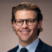 John, a white man with brown hair and circular brown glasses, wears a pale blue shirt, black jacket, and dark blue tie. 