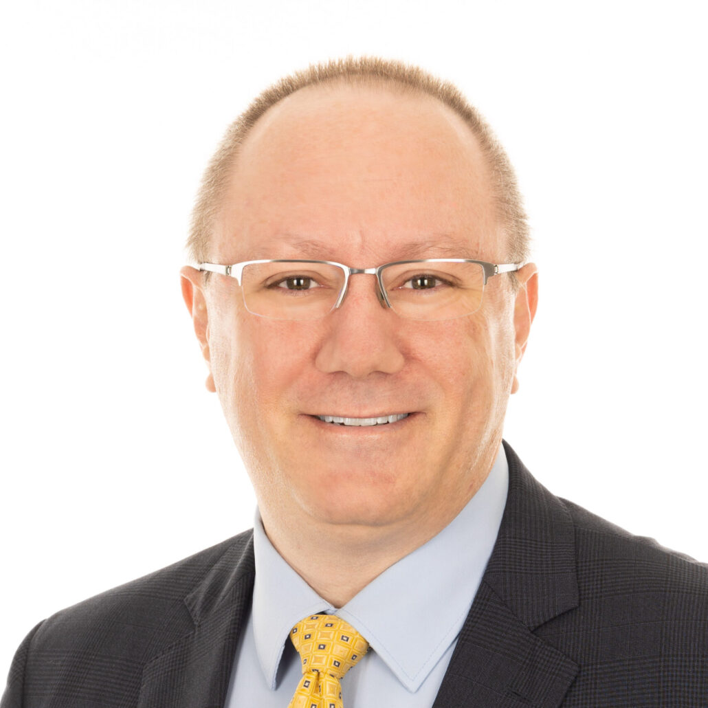 Reed, a white man with light brown hair, wears clear glasses, a pale blue shirt, a black jacket, and a yellow tie with blue and white squares.