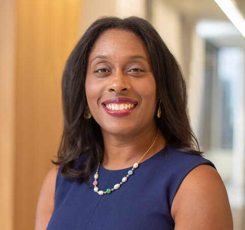 Shana, a Black woman with dark brown hair, wears a navy blue dress and a red, blue, and yellow beaded necklace.