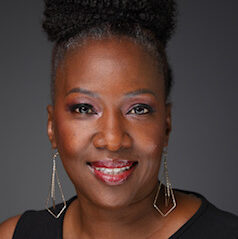 Lakisha, a Black woman with black hair in a bun, wears a black sleeveless shirt and gold earrings.