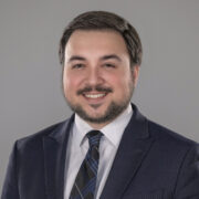 Christian, a white man with brown hair, wears a white shirt, dark blue tie, and black suit.