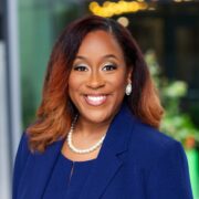 Chazle, a Black woman with black hair and auburn highlights, wears a blue suit and blazer and a pearl necklace.