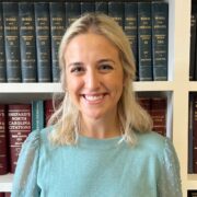 Alexa Lyles Brinegar is a woman with blond hair. She wears teal blouse and is standing in front of a bookshelf with legal books.