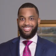Jay, a Black man with black hair and a beard, wears a white shirt, pink tie, and navy jacket.