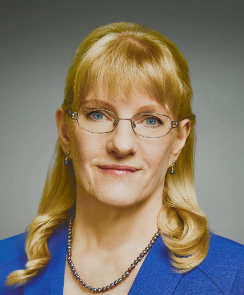 Lauren, a white woman with blond hair and bangs, wears wire-rimmed glasses, a royal blue blouse and gold necklace.