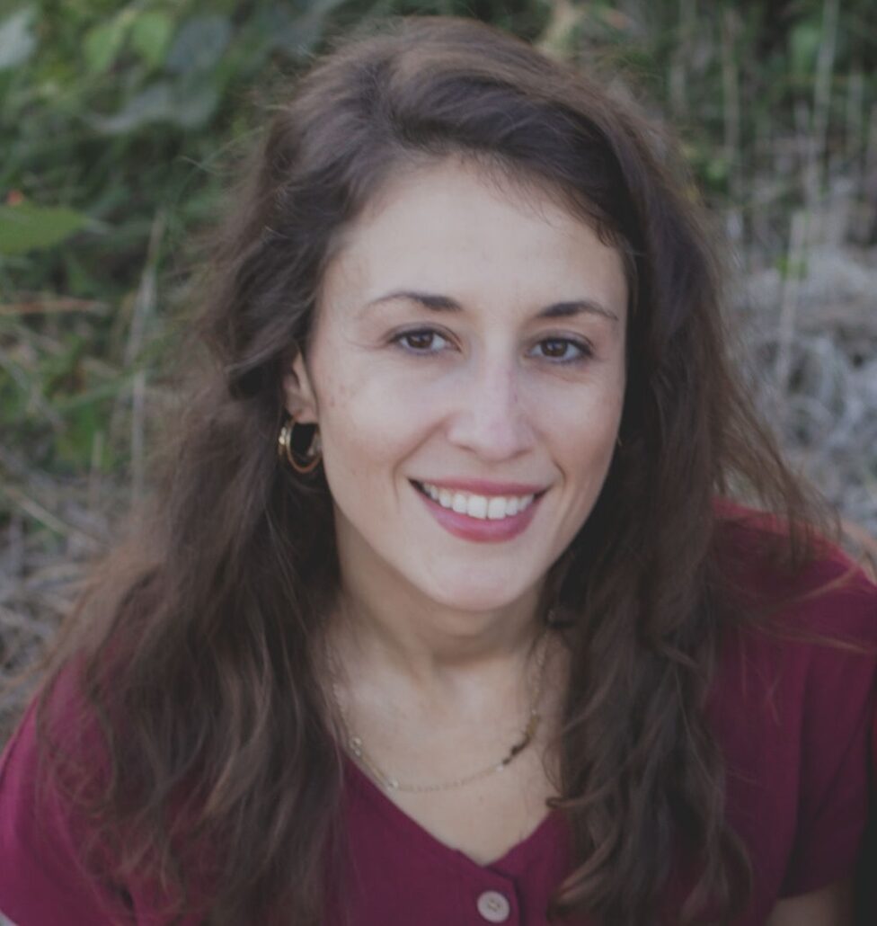 Lauren, a white woman with brown hair, wears a burgundy colored blouse. Foliage in the background.